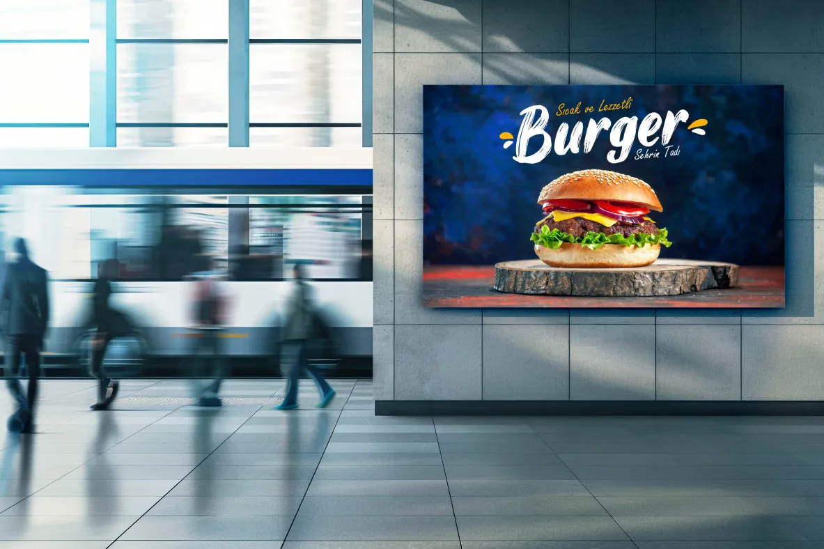 Modern bir transit istasyonunun içindeki bir duvarda marul, soğan ve domatesli bir burger gösteren büyük bir burger reklamı. Arka planda bulanık bir tren ve insanlar görülüyor.