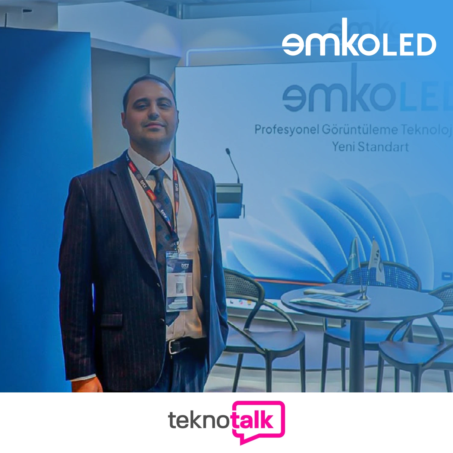 Takım elbiseli bir adam, LED Teknolojisi'ni sergileyen EmkoLED'in teknoloji fuarındaki standında gülümseyerek duruyor. Mavi marka ve boş sandalyeler onu çevrelerken altta teknotalk yazısı beliriyor.